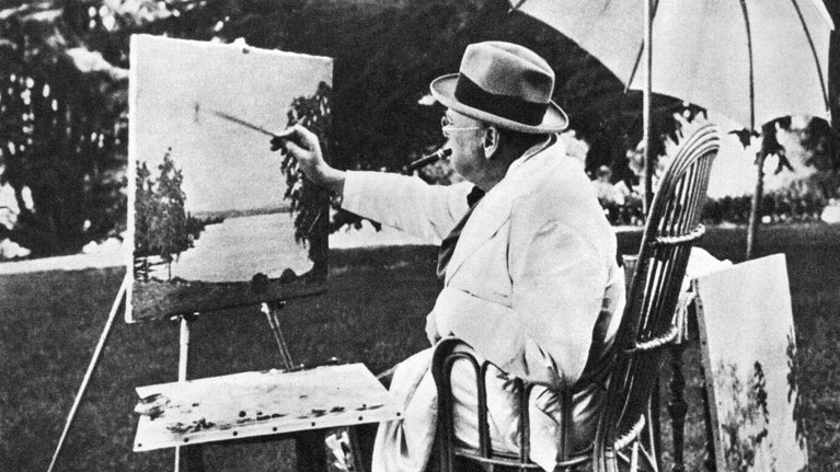 Black and white archive photo of Sir Winston Churchill seated before an easel and paint set painting beneath a parasol.